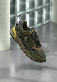 Olivengrønn sneaker med svarte detaljer, brun semsket skinn og reflekterende logo. Har teksturert mesh og polstret såle for støtte.