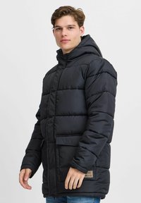 Chaqueta de plumas negra con capucha, textura acolchada, bolsillos delanteros y una etiqueta de marca cerca del dobladillo. Presenta un cierre con cremallera y mangas largas.