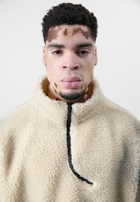 Fleece-Pullover in Creme mit strukturiertem Material, hohem Kragen und schwarzem Reißverschlussdetail. Das Innere hat eine weiche, plüschige Textur.