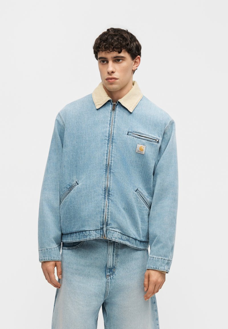 Jonge man draagt een lichtblauwe spijkerjas met beige kraag en bijpassende lichtblauwe spijkerbroek tegen een effen lichte achtergrond.