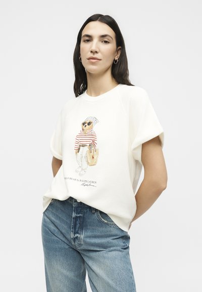 Γυναίκα με μακριά σκούρα μαλλιά που φοράει λευκό κοντομάνικο πουκάμισο με σχέδιο σκύλου με γυαλιά ηλίου και μπλε τζιν, στέκεται μπροστά σε απλό φόντο.