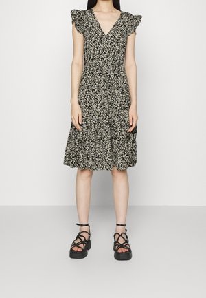 Kobieta w czarnej, kwiatowej sukience sięgającej do kolan z falbankowymi rękawami i czarnych sandałach na platformie z paskami wokół kostek, stoi na tle jednolitego tła.