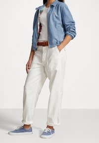 Jasnoniebieska bomberka z denimu noszona na białej koszulce, przewiązana plecionym brązowym paskiem. Białe dopasowane spodnie oraz niebieskie trampki dopełniają stylizację.