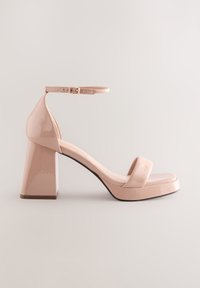 Sandale à talon haut en cuir verni couleur nude, avec une large bride sur les orteils, une bride de cheville ajustable, et un talon rectangulaire épais.