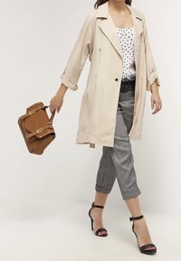 Cappotto beige con grandi risvolti, design doppiopetto, abbinato a un top a pois bianco e nero, pantaloni grigi alla caviglia e décolleté nere. Borsa marrone.