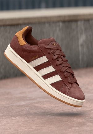 Bruine suède sneaker met witte strepen en gumzool, merk Campus, getoond in de lucht tegen een getextureerde grijze en roze achtergrond.