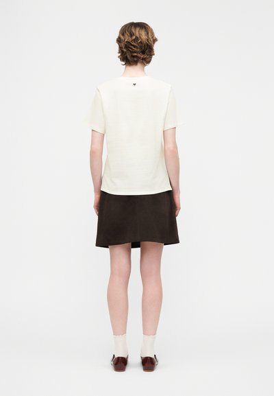 Άτομο με κοντά, κυματιστά μαλλιά στέκεται γυρισμένο με την πλάτη, φοράει λευκό μπλουζάκι με κοντά μανίκια, μαύρη φούστα, λευκές κάλτσες και καφέ παπούτσια, σε λευκό φόντο.