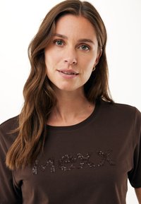 Brązowy bawełniany t-shirt z okrągłym dekoltem i krótkimi rękawami, ozdobiony błyszczącymi cekinami, które tworzą napis "Mexx" na przodzie.