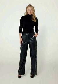 Kvinna med blont hår som bär en svart polotröja och svarta vida läderbyxor med fickor, stående med en hand i fickan.