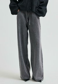 Persona, kura uzvilkusi augstās jostasvietas, plašās, izbalējušās melnās džinsās un melnu pogātā kardigānā ar rokām kabatās, stāv uz gaiša fona.