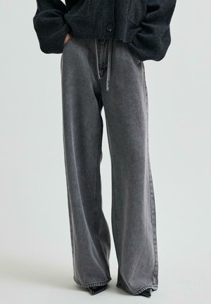 Persoon in een hoog-getailleerde, wijde, vervaagde zwarte spijkerbroek en een zwarte cardigan met knopen, met handen in de zakken, staand tegen een lichte achtergrond.