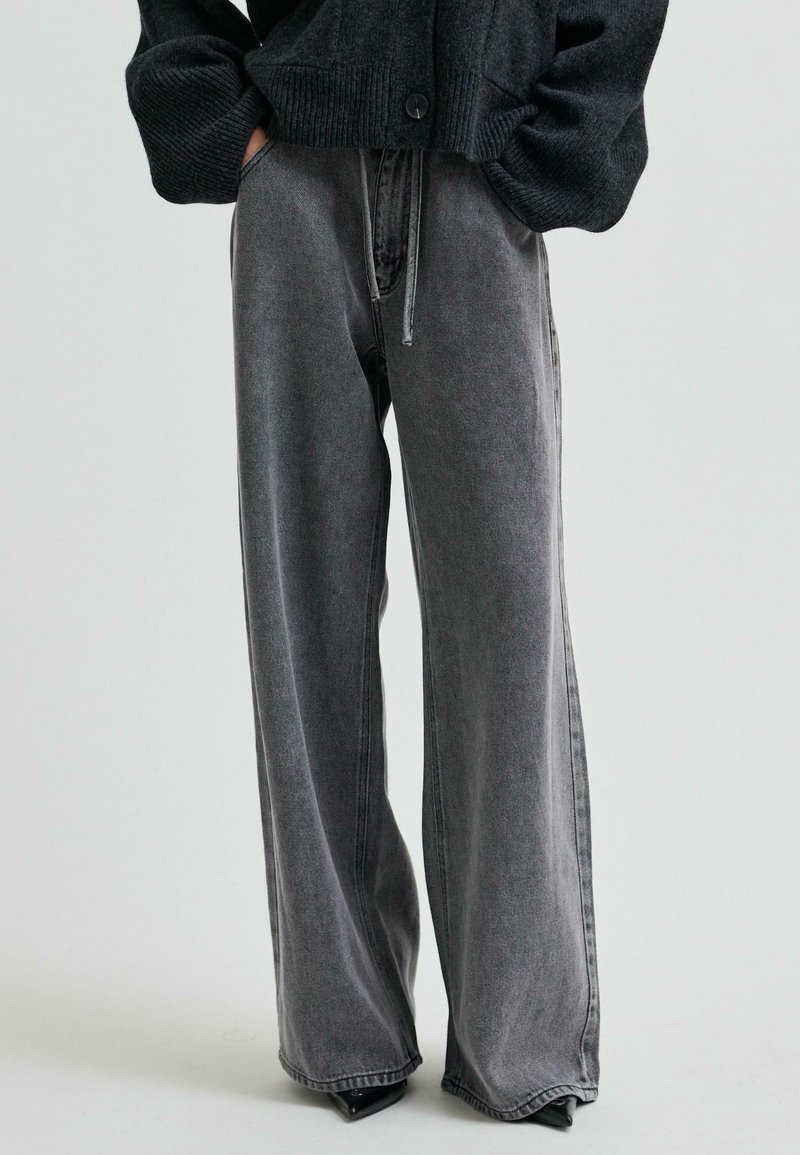 Persona, kura uzvilkusi augstās jostasvietas, plašās, izbalējušās melnās džinsās un melnu pogātā kardigānā ar rokām kabatās, stāv uz gaiša fona.