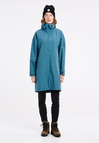Tyrkysová nepremokavá parka s vysokým golierom, zipsovým zapínaním a nastaviteľnou kapucňou. Nosená cez čierne nohavice a hnedé turistické topánky.