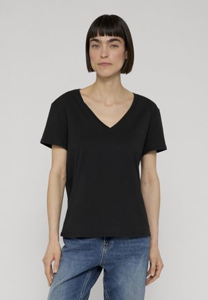 Donna con capelli corti e scuri, indossa una maglietta nera a V e jeans blu, in piedi davanti a uno sfondo grigio chiaro uniforme.