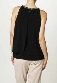 Svart ärmlös topp med rundad halsringning, med detaljer av pärlor vid halsringningen och plissér på baksidan, tillverkad av lättviktsmaterial. Matchas med ljusa byxor.