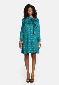 Turkusowa sukienka z geometrycznym żółtym wzorem, długimi rękawami, związanym dekoltem i warstwowym dołem. Miękka tkanina i luźny krój.