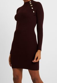 Robe en maille côtelée bordeaux avec un col montant, des manches longues et des accents de boutons dorés sur l'épaule. Silhouette ajustée.