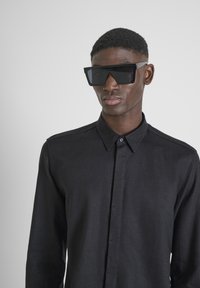 Camicia nera con bottoni, dalla texture liscia e con colletto; indossata con grandi occhiali da sole neri, angolari. Sfondo semplice.