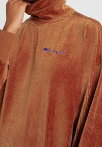 Col roulé en velours côtelé marron présentant une texture côtelée, une coupe oversize et un logo "Champion" brodé en bleu sur la poitrine.