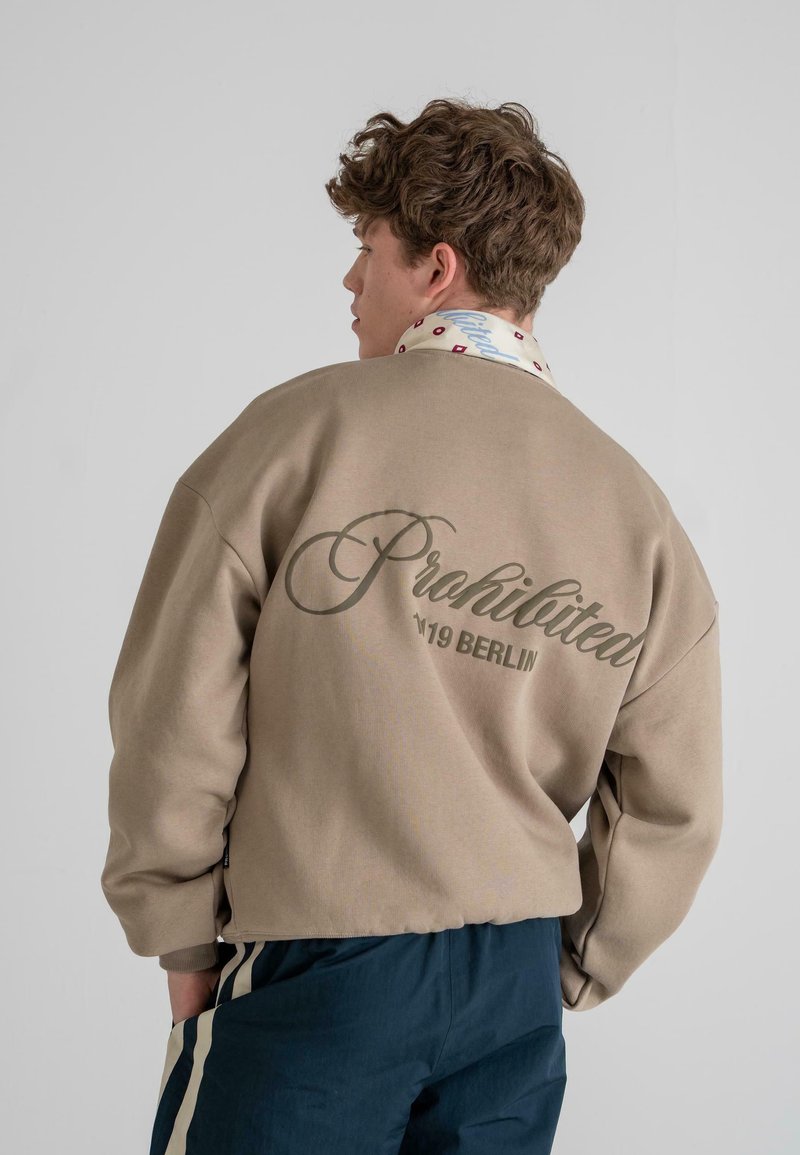 Beige sweatshirt with oversized fit, featuring cursive "Prohibited" text and "19 Berlin" printed on the back. Accompanied by a patterned shirt collar.