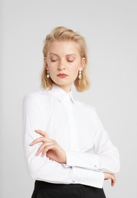 Chemise blanche à manches longues avec col pointu et poignets à bouton, ornée de détails métalliques sur les poignets. Tissu en coton lisse.