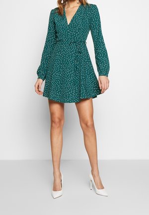 Vestido azul verdoso con estampado de lunares, diseño envolvente, mangas largas y cintura con lazo. Un par de zapatos de tacón blanco completa el look.