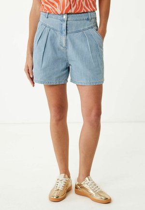 Shorts in denim bluastro con pieghe, due tasche anteriori e chiusura con bottone. Abbinati a sneakers dorate lucide e a una maglietta arancione con motivo.