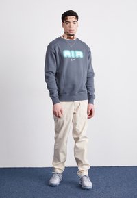 Dark grey sweatshirt features a large light blue "AIR" logo with a Nike swoosh. Paired with beige cargo pants and grey sneakers.