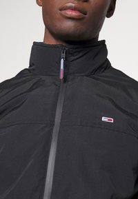 Närbild av en person som bär en svart zip-up jacka med en liten röd, vit och blå logotyp på bröstet och dragkedjans grepp.