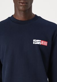 Mørkeblå hettegenser laget av bomull med ribbet rund halslinning. Har en hvit logo og røde detaljer på brystområdet.