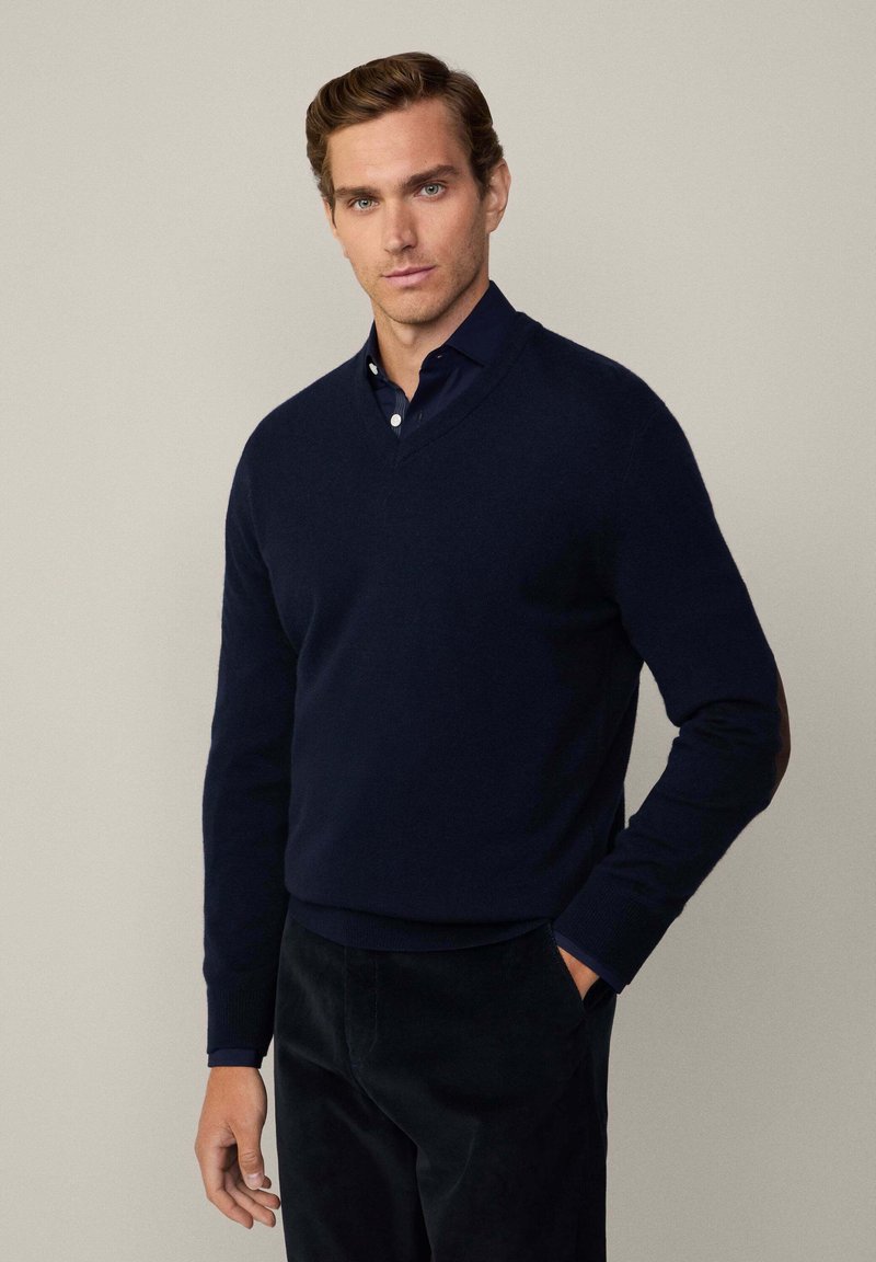 Tummansininen, V-pääntie neulepaita pehmeästä neuloksesta ribatulla hihalla. Pukeutunut kauluspainan päälle, yhdistetty tummiin housuihin.