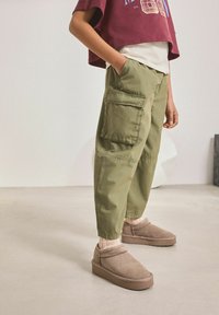 Cargo byxor i olivgrön tyg med sidofickor, kombinerat med bruna slip-on skor och beige strumpor, bärandes en kortärmad maroon topp.