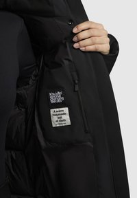 Fekete dzseki sima felülettel, belső címkével és cipzáras zsebbel. Puha bélés és visszafogott fém részletek láthatók.