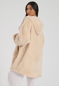 Beige gewatteerde pluche jas met een zachte, gestructureerde afwerking. Heeft een losse pasvorm en grote zakken, en reikt tot halverwege de bovenbenen.