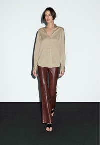 Camicia beige con bottoni e colletto, maniche lunghe, abbinata a pantaloni in pelle marrone a campana e sandali neri senza cinturino su uno sfondo bianco.