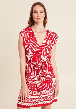 Frau in ärmellosem, rot-weiß gemustertem Kleid mit Blättern, V-Ausschnitt und gebundener Taille, steht vor einem schlichten beigefarbenen Hintergrund.