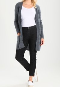 Grå lång kofta i mjukt tyg med ribbade ärmar, tillsammans med svarta slim-fit jeans och vita espadrilloskor. Neutral bakgrund.