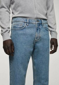 Jeans in denim azzurro chiaro con una vestibilità classica, dotati di cinque tasche, cuciture tono su tono e gamba dritta, abbinati a una camicia grigia a maniche lunghe.