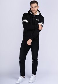 Čierna mikina na zips s umelou kožušinou, biele pruhy na rukávoch, klokanie vrecká, skombinované s čiernymi džínsami a bielymi teniskami.