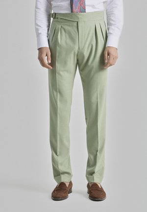 Pantalones plisados de color verde claro, hechos de una tela suave, con una cinturilla ancha y un hebilla lateral. Combinados con zapatos marrones.