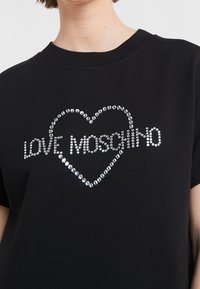 Černé tričko s designem srdce z stříbrných kamínků, s textem "LOVE MOSCHINO" v dekorativním písmu. Krátké rukávy a kulatý výstřih.