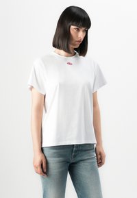 Kvinne med rett svart hår som har på seg en enkel hvit T-skjorte med en liten rosa logo og lyseblå jeans, stående mot en hvit bakgrunn.