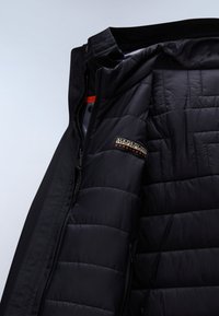 Sort pufferjakke med quiltet tekstur, der har lynlås i lukning og en orange detalje. Logo-patch på venstre brystområde.