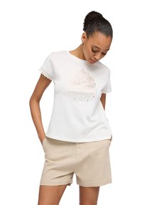 Biała bawełniana koszulka z koronkowym wykończeniem, z różowo-złotym wzorem lotosu na przodzie. W połączeniu z beżowymi szortami, modelka przyjmuje swobodną pozę.