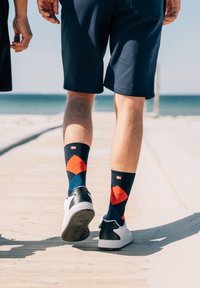 Bomuldsblandede sokker med en marineblå baggrund, prydet med røde og orange diamantmønstre. Båret med sorte og hvide sneakers på en strandpromenade.