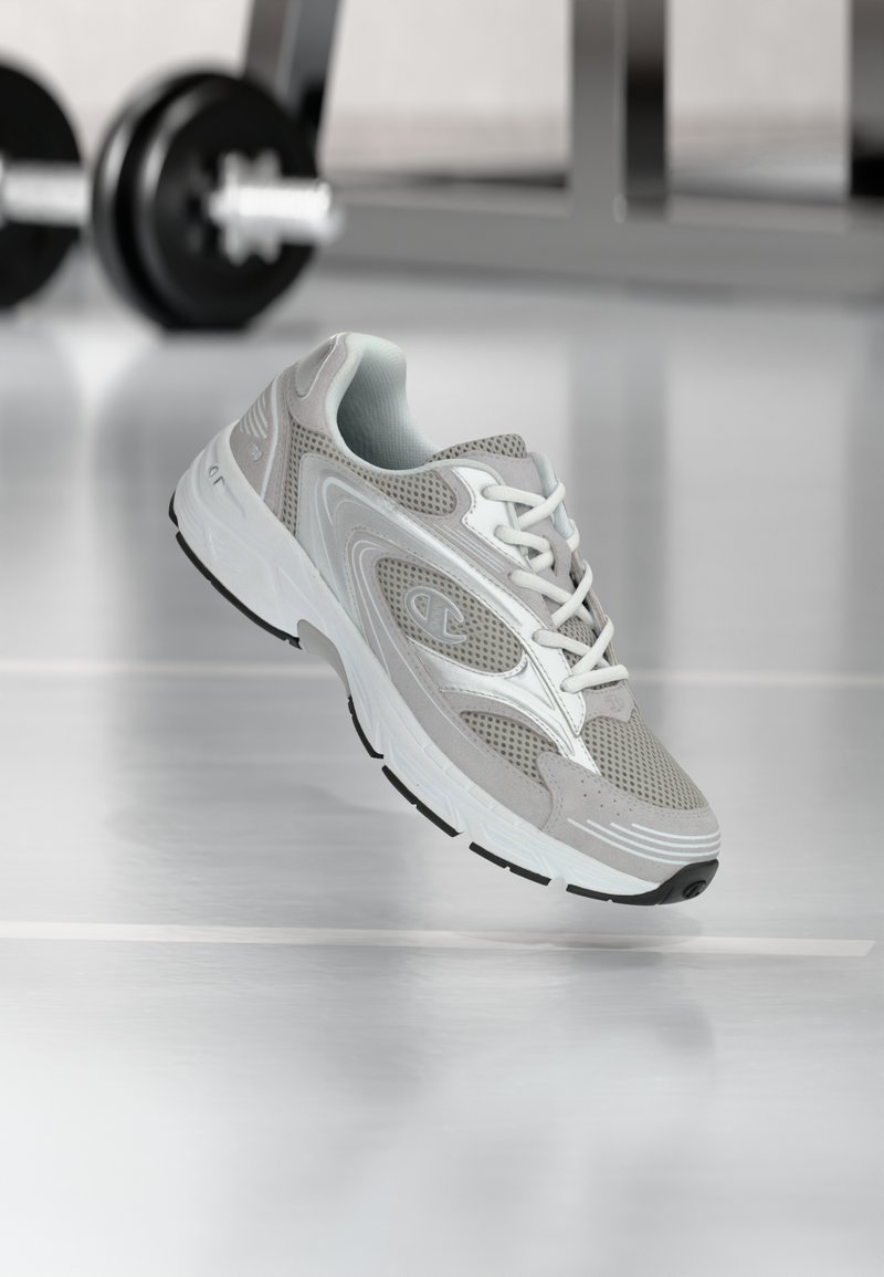 Αθλητικό παπούτσι ανοιχτού γκρι με επάνω μέρος από δίχτυ και σουέτ, λευκά κορδόνια, μαύρη σόλα και καμπυλωτό σχέδιο· διαθέτει λογότυπο στο πλάι.