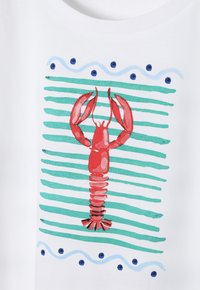 Weißes T-Shirt mit einem roten Hummer-Motiv, das auf einem gestreiften grünen Hintergrund mit blauen Punkten und wellenförmigen Linien zentriert ist.