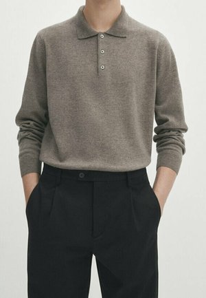 Person wearing a taupe long-sleeve polo shirt tucked into high-waisted black tailored trousers with hands in pockets.