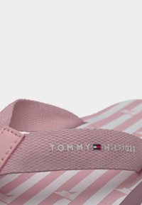 Ροζ λουράκι σαγιονάρας με υφή και λογότυπο "TOMMY HILFIGER" μαζί με τη σημαία, τοποθετημένο σε ροζ και λευκή σανδάλι με σχέδια αστεριών.