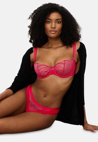 Γυναίκα με σγουρά μαλλιά φορεμένη με κόκκινο σουτιέν δαντέλα και ταιριαστά ε�σώρουχα, μαύρος κάρντιγκ ριγμένος στους ώμους, καθισμένη σε λευκό φόντο.